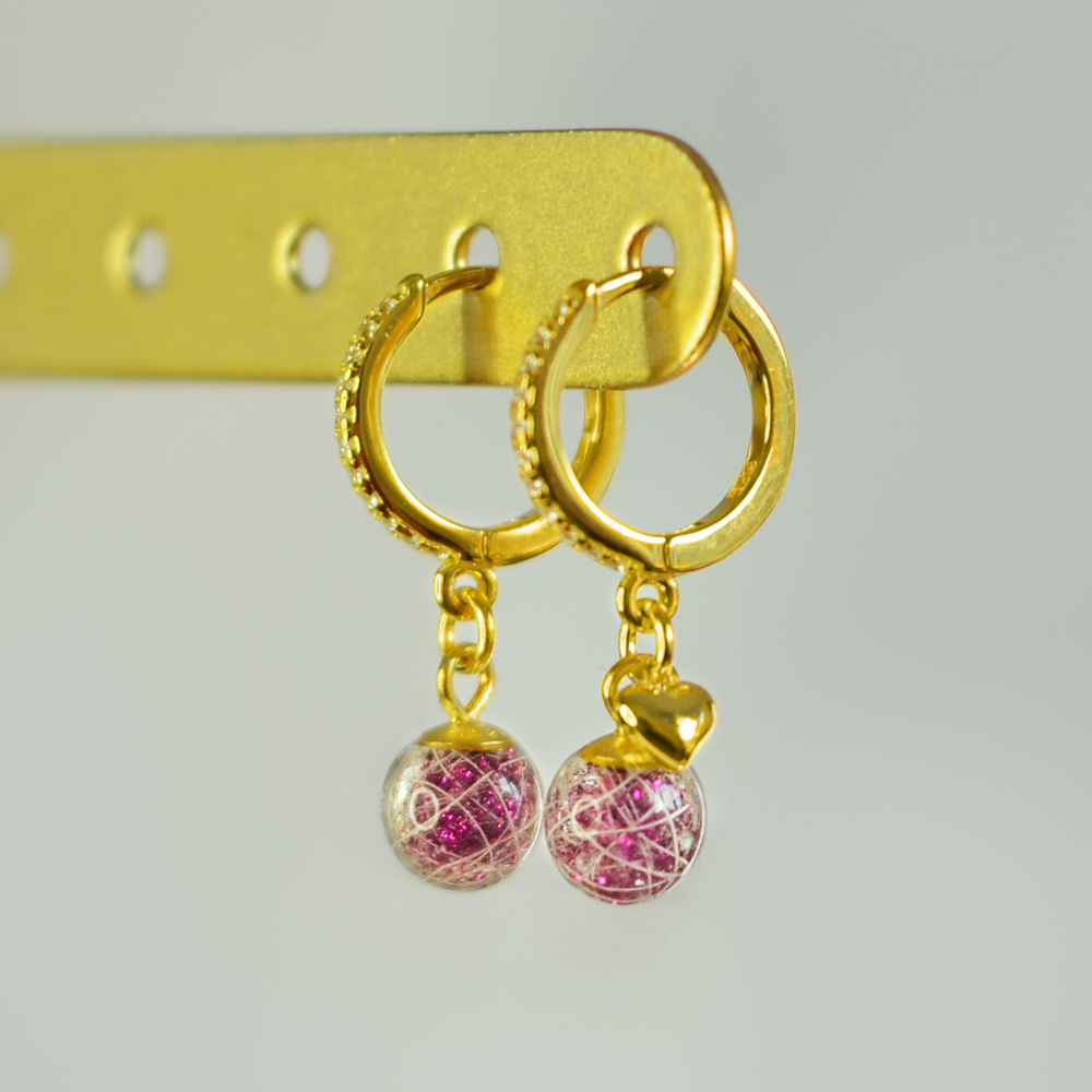 Lusia øreringe i forgyldt 925 sterling sølv med rhinestones og to håndlavede resinperler med hår fra palomino pony. Lyserødt glimmer i perlen fremhæver de hvide hår. Set fra siden med fokus på øreringen med minihjerte. I firkantet format.